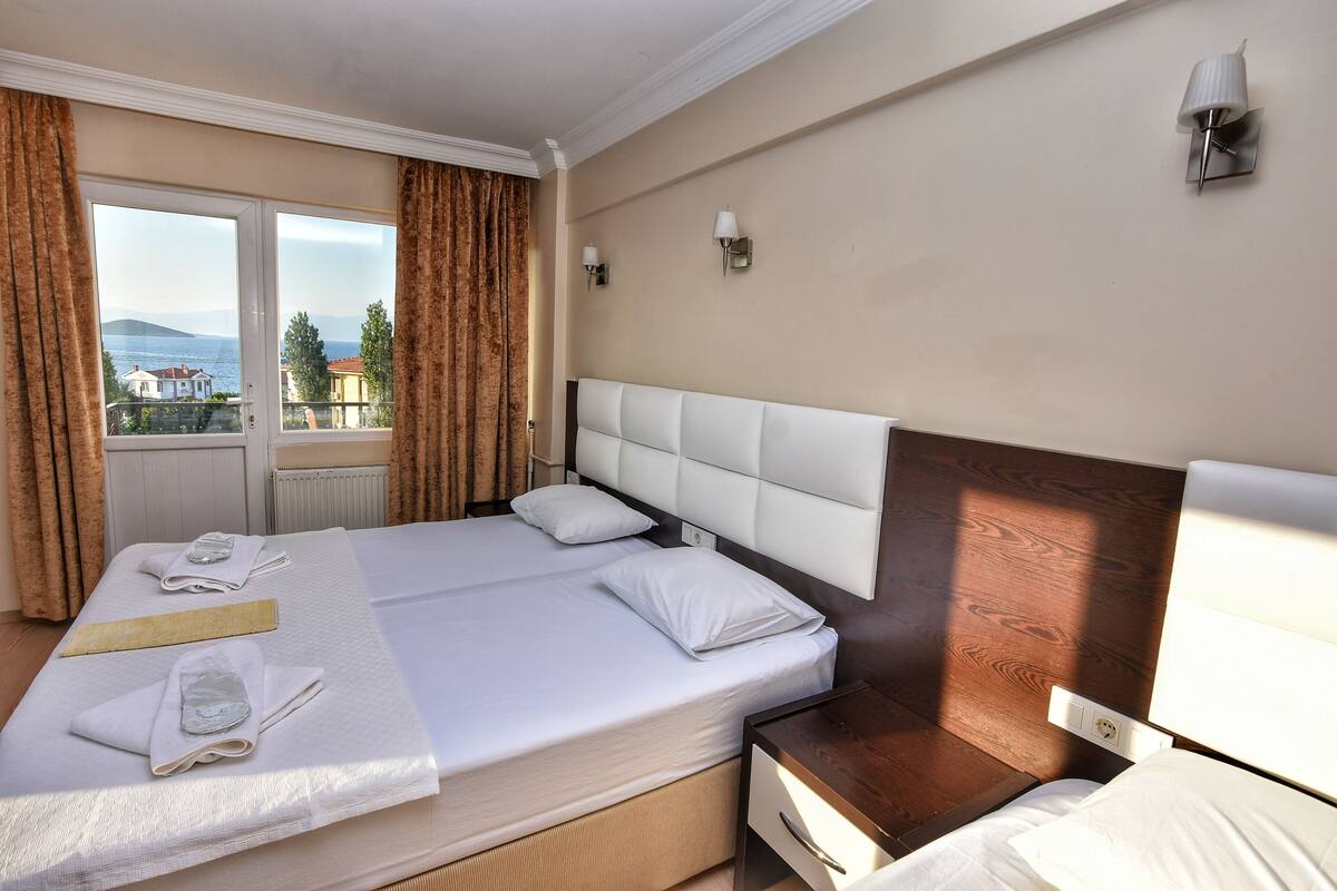 Une chambre d’hôtel avec un grand lit, une tête de lit en bois, deux tables de chevet et une vue sur la mer par une fenêtre agrémentée de rideaux.