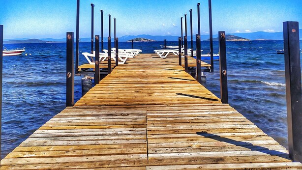 A wooden pier extending into the water with white lounge chairs and a boat in the background. - Uzunhan Hotel (Ayvalik)