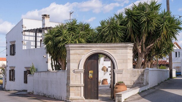 A white building with a stone archway entrance, palm trees, and a tiled roof. - Villa Tzivaeri (Chania)
