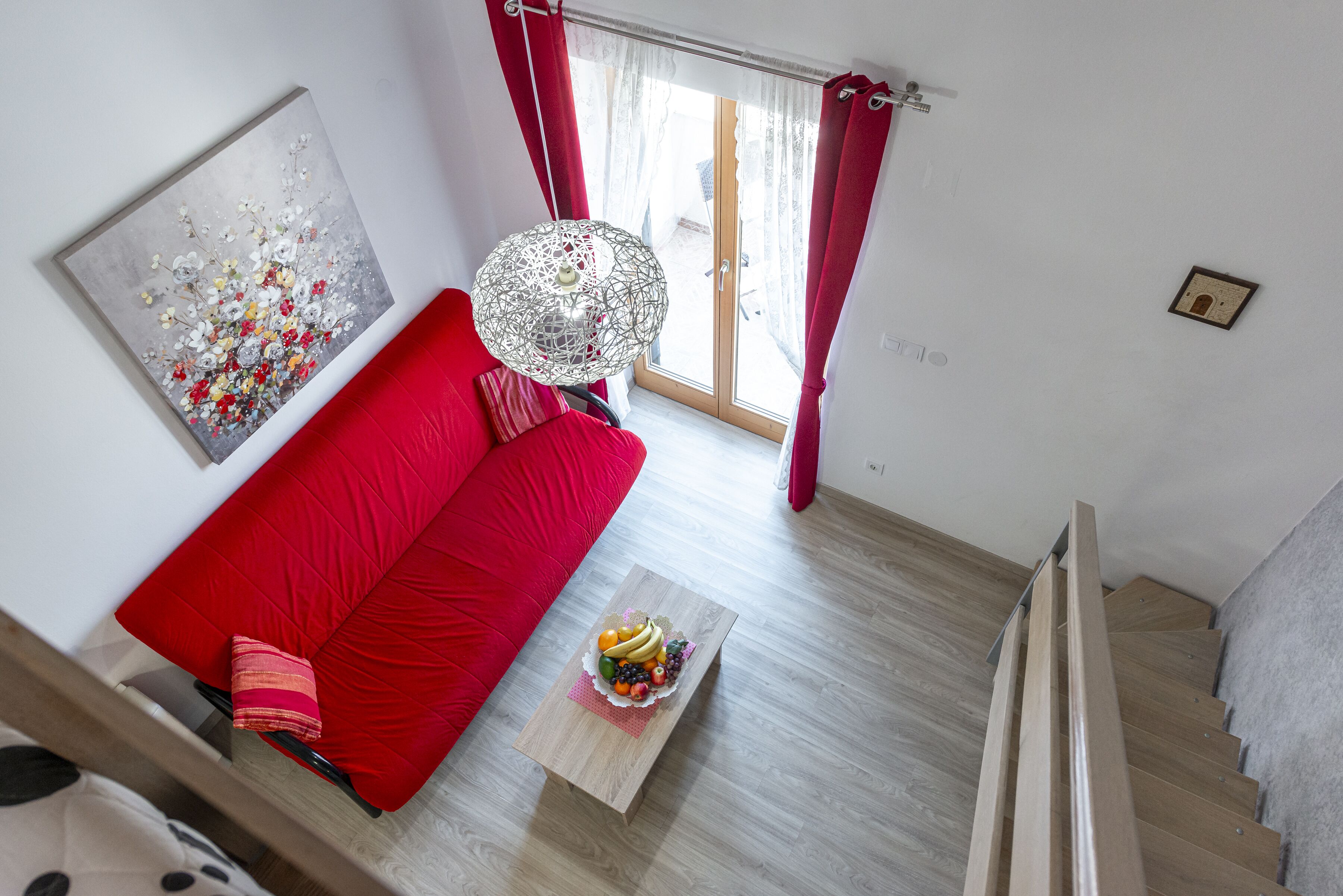 Una sala de estar moderna con un sofá rojo, una mesa de centro de madera con fruta, un cuadro abstracto grande y vistas al exterior a través de una puerta de cristal.
