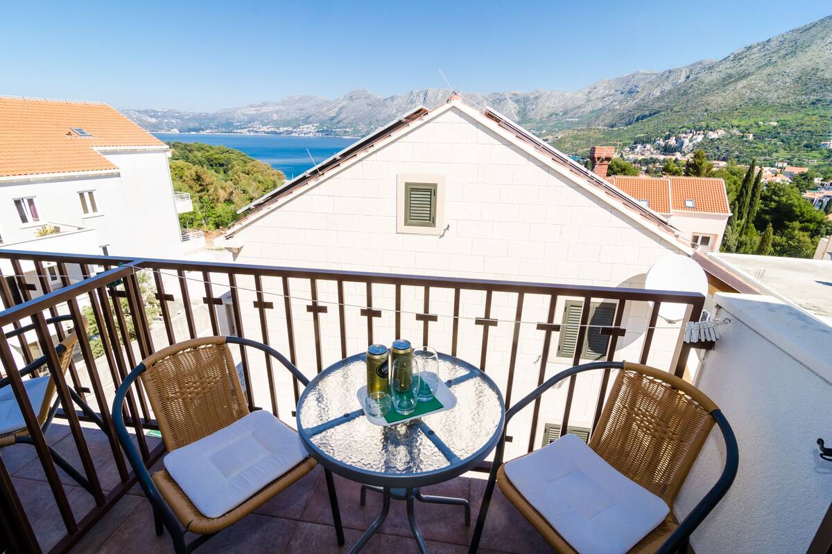 A balcony with wicker chairs and a glass table, overlooking a mountainous landscape and the sea.