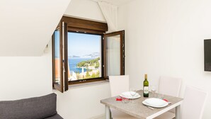 A dining area with a table set for two, a view of the sea, and a window with curtains. - Cina Apartments (Konavle)