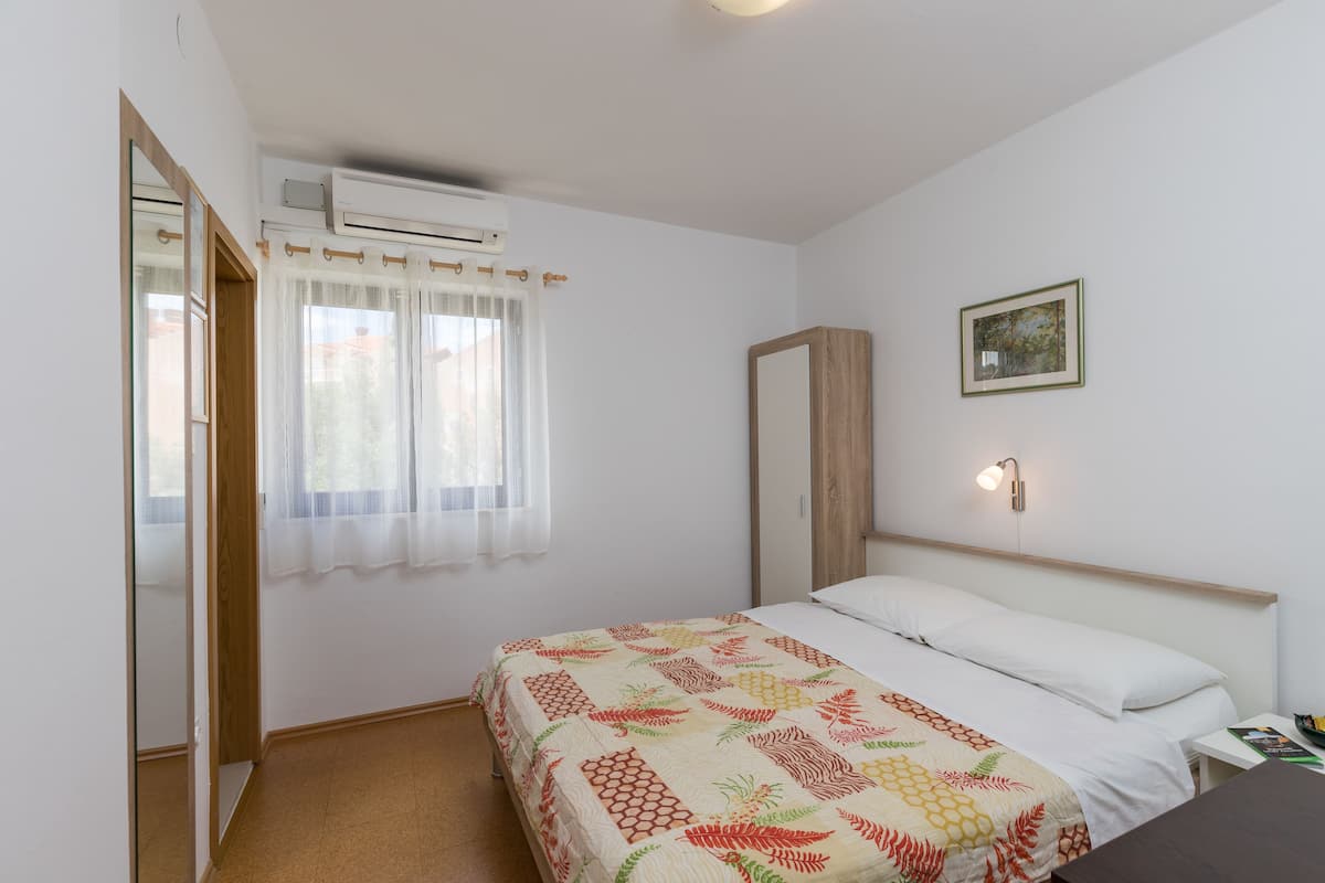 A bedroom with a bed, a bedside table, a nightstand, a mirror, a window with curtains, an air conditioning unit, and a framed picture on the wall.
