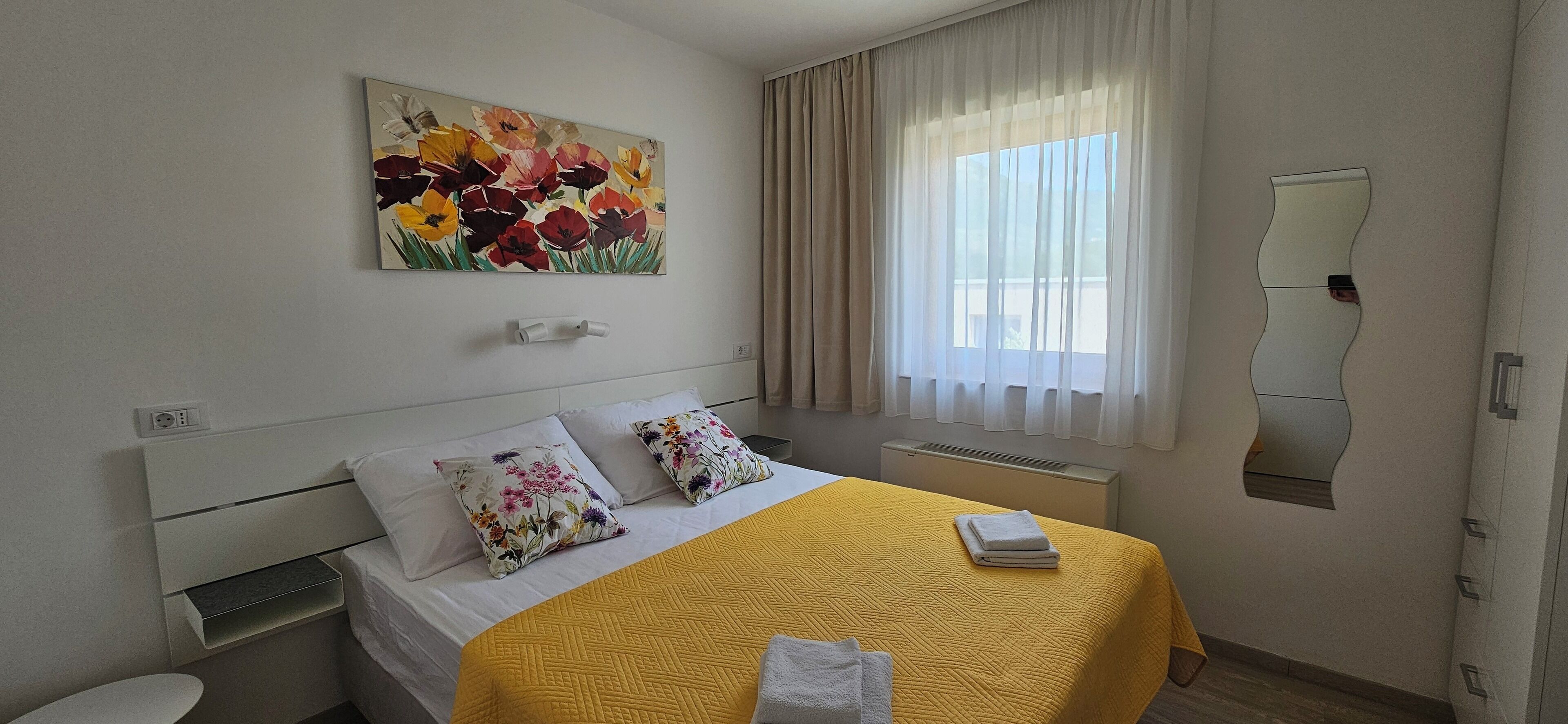 Quarto de hotel com uma cama, um quadro de flores na parede, uma janela com cortinas e um espelho.