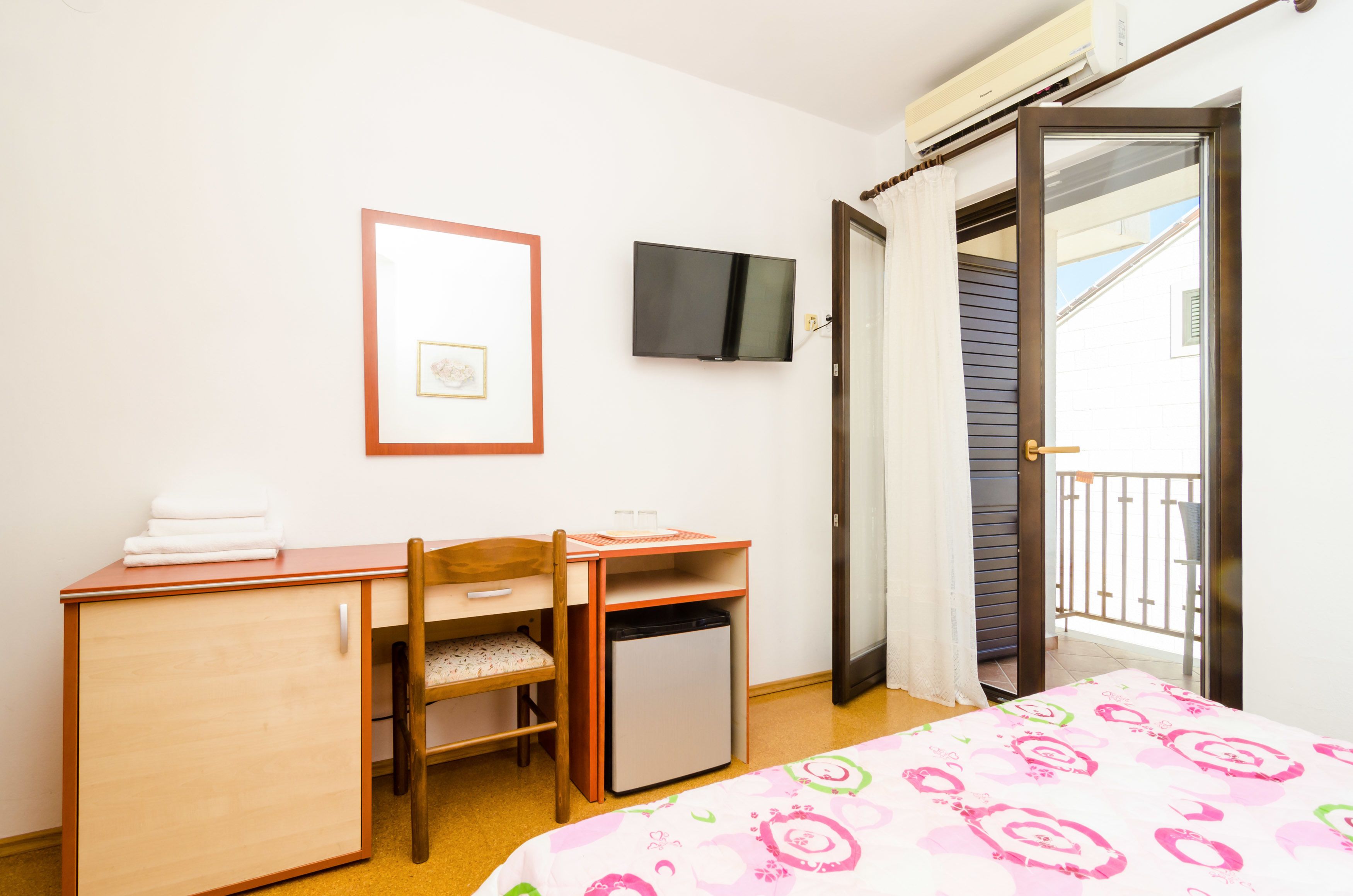 A bedroom with a bed, a desk, a chair, a small refrigerator, a TV mounted on the wall, and a balcony door.