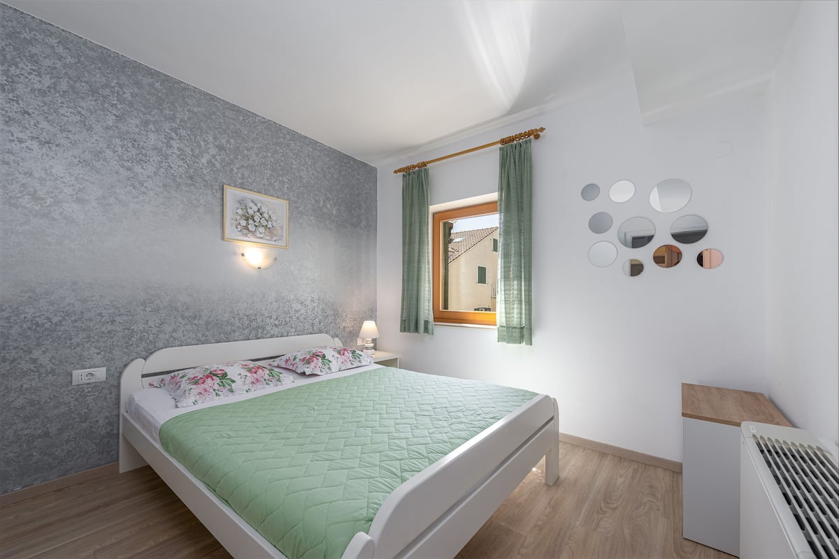 A bedroom with a bed, a bedside table, a wall-mounted light, a window with curtains, and a radiator.