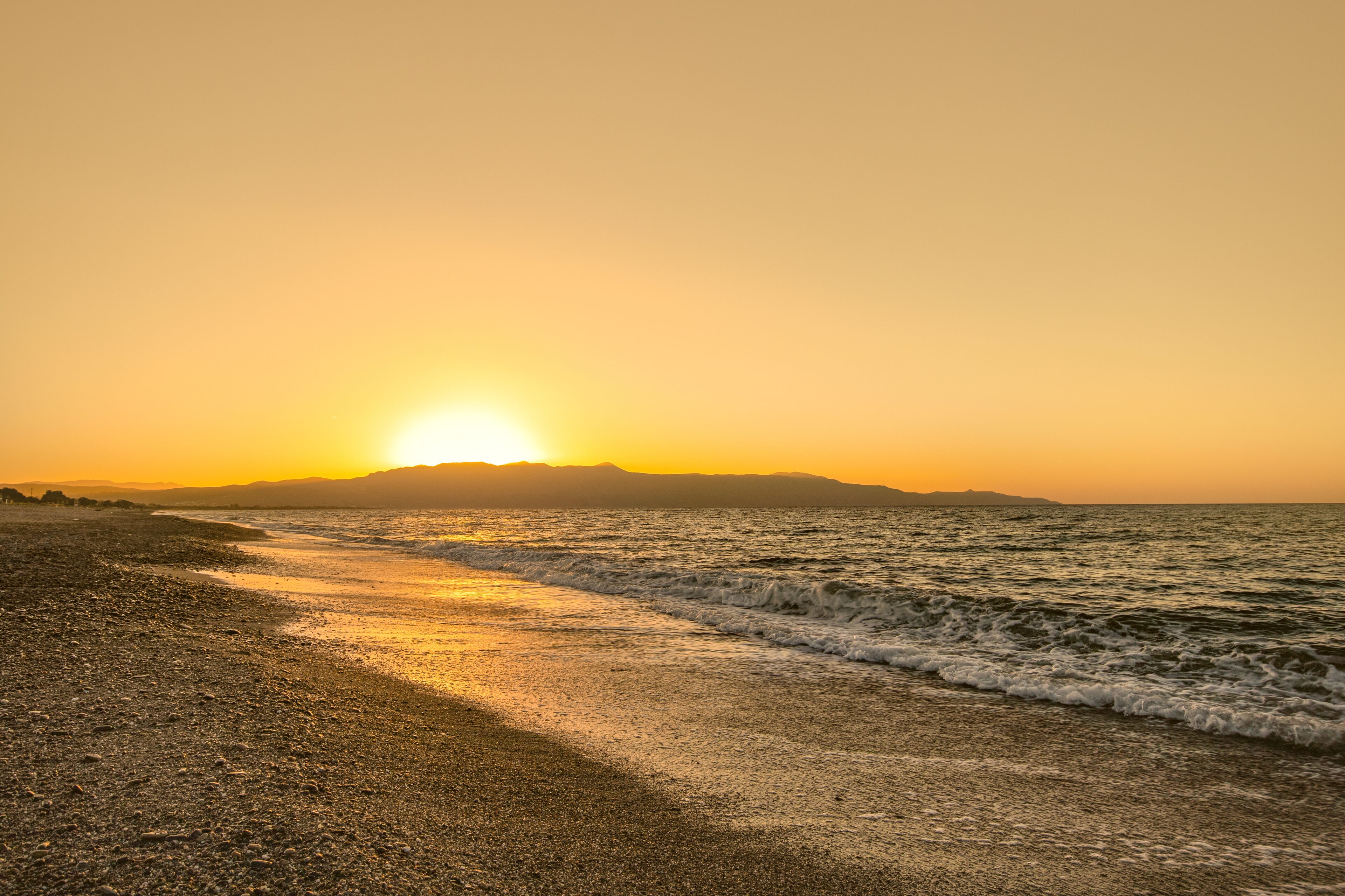 Una playa con atardecer, olas y montañas a lo lejos.