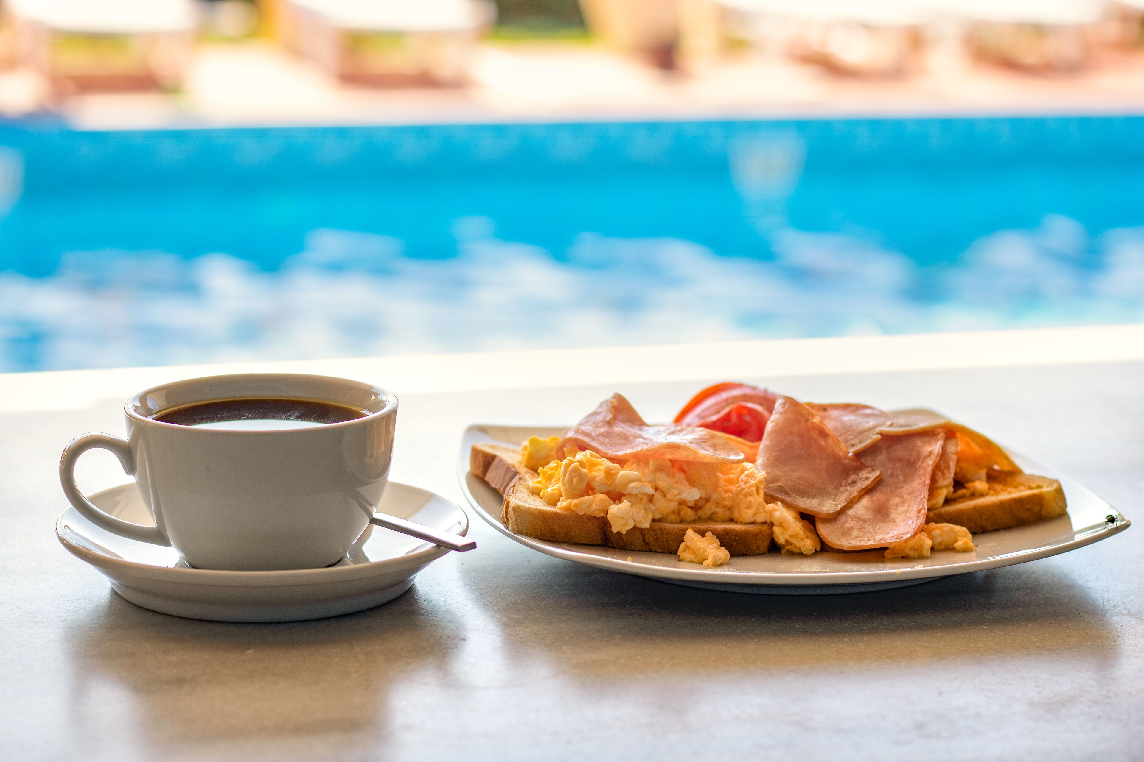 Un plato de desayuno con huevos revueltos, jamón y tostadas, acompañado de una taza de café, sobre una mesa con vista a una piscina.