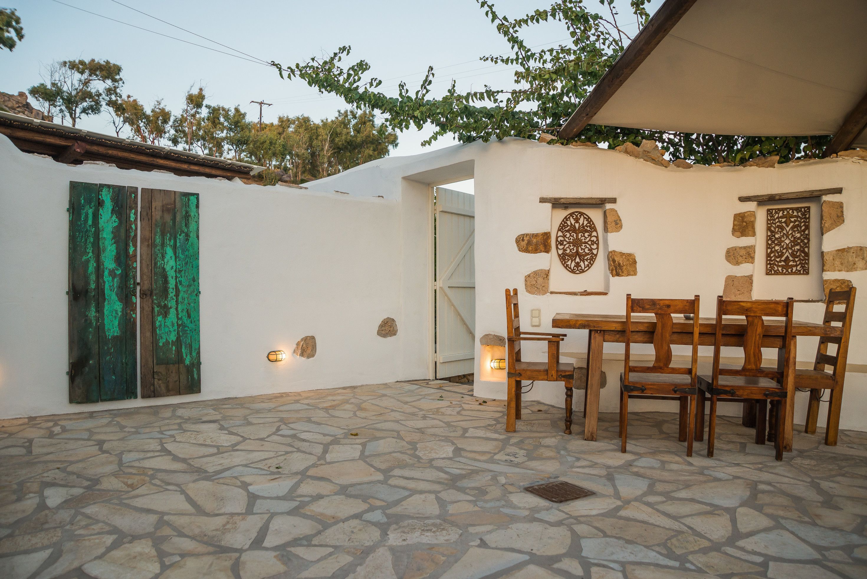 A stone-floored patio with a wooden table and chairs, a stone wall with decorative elements, and a door with green shutters.