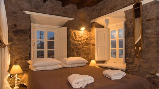 Une chambre aux murs de pierre, avec un lit, deux fenêtres, un miroir et une lampe de chevet.