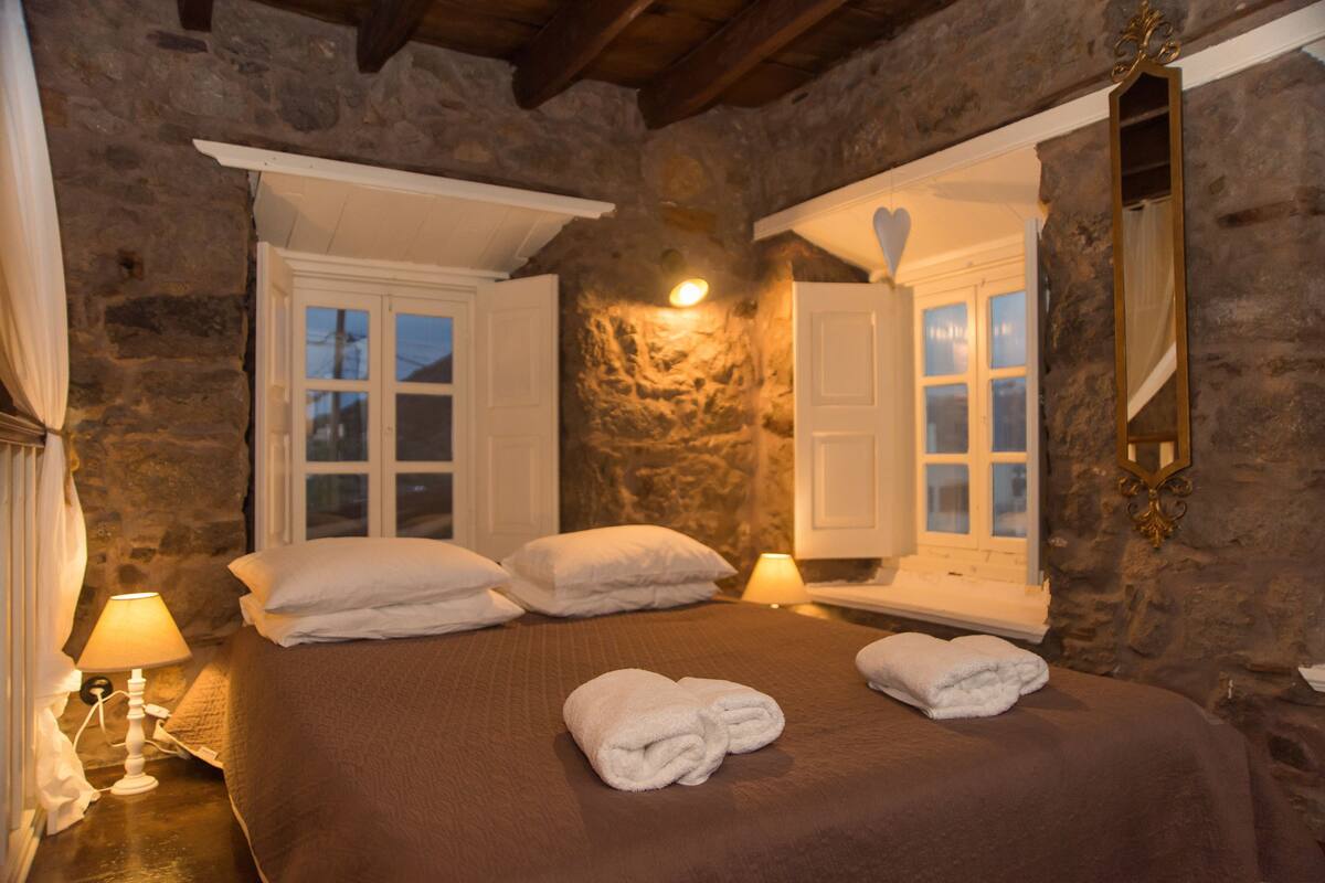 A stone-walled bedroom with a bed, two windows, a mirror, and a bedside lamp.