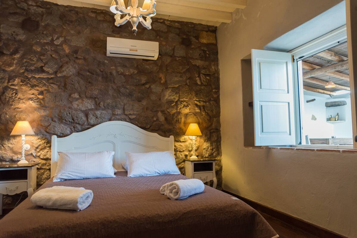 A bedroom with a stone wall, a white bed, two bedside lamps, and a window with open shutters.
