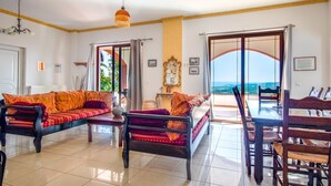 A spacious living room with a sofa, dining table, and chairs. There is a view of the ocean through large windows.