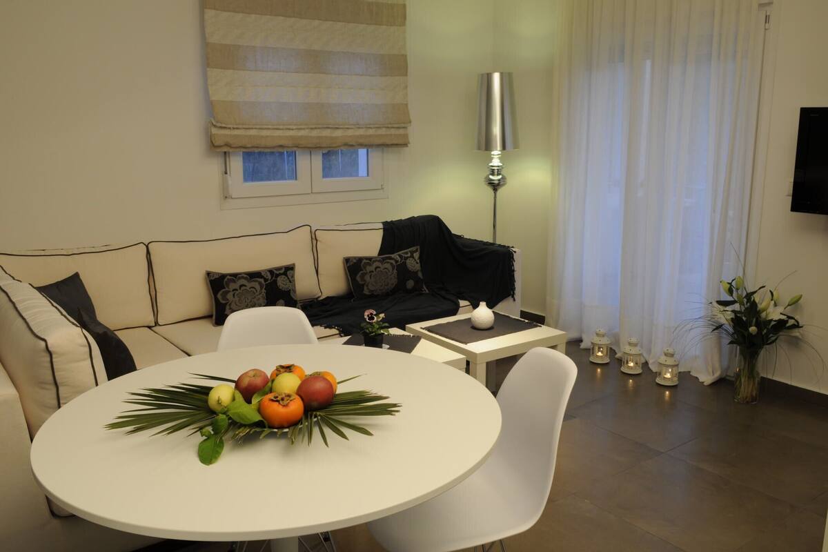 A living room with a sofa, a round table with fruit, a lamp, and a TV on the wall.