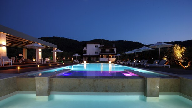 Une piscine entourée de chaises longues et de parasols, bordée d’un immeuble à plusieurs étages et avec une chaîne de montagnes en arrière-plan.