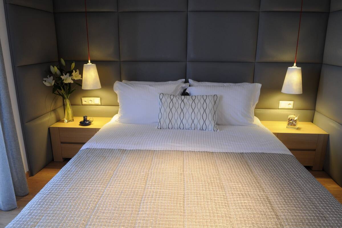A neatly made bed with white linens, two bedside lamps, a vase with flowers, and a wall-mounted light fixture.