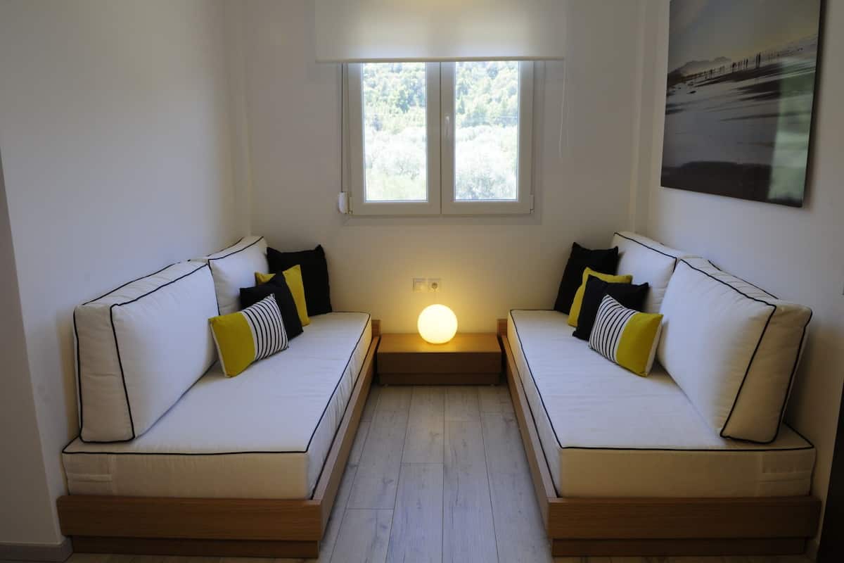 A room with two white sofas with black and yellow cushions, a wooden floor, a window with a view of trees, and a wall-mounted light fixture.
