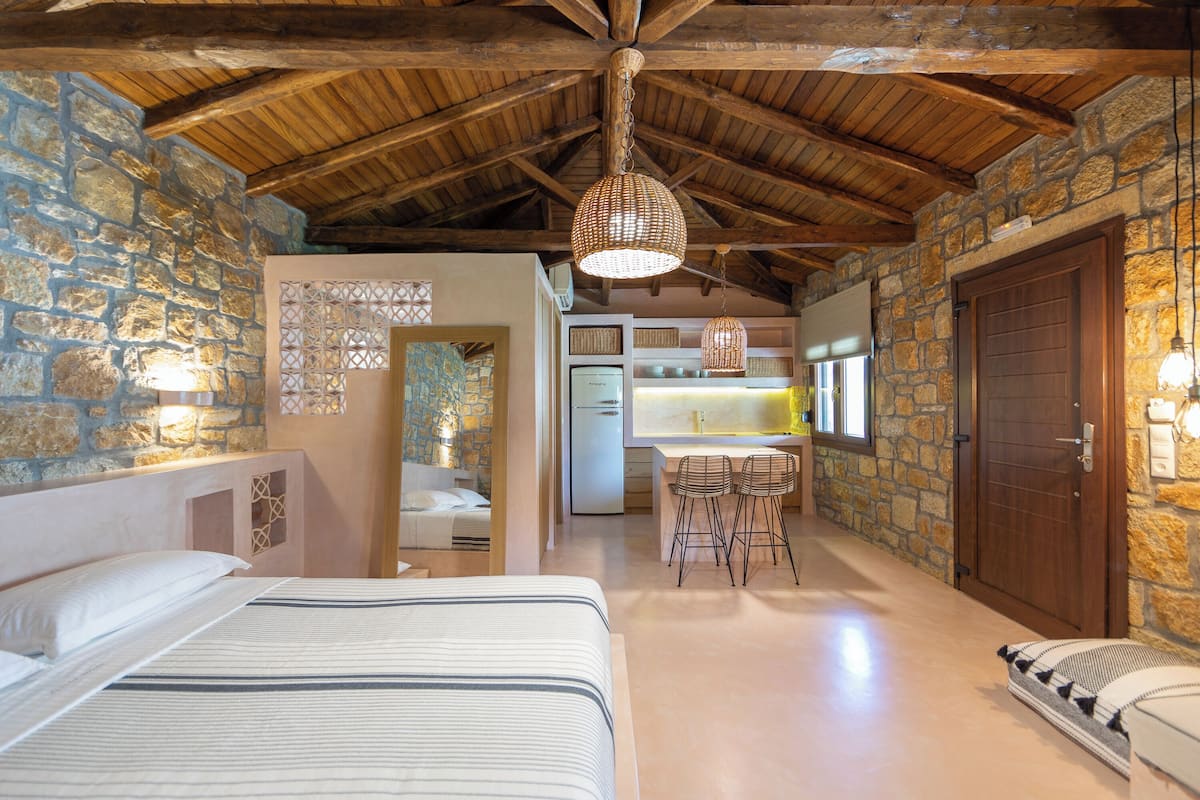 A cozy room with a bed, a desk with chairs, a stone wall, and a wooden ceiling.