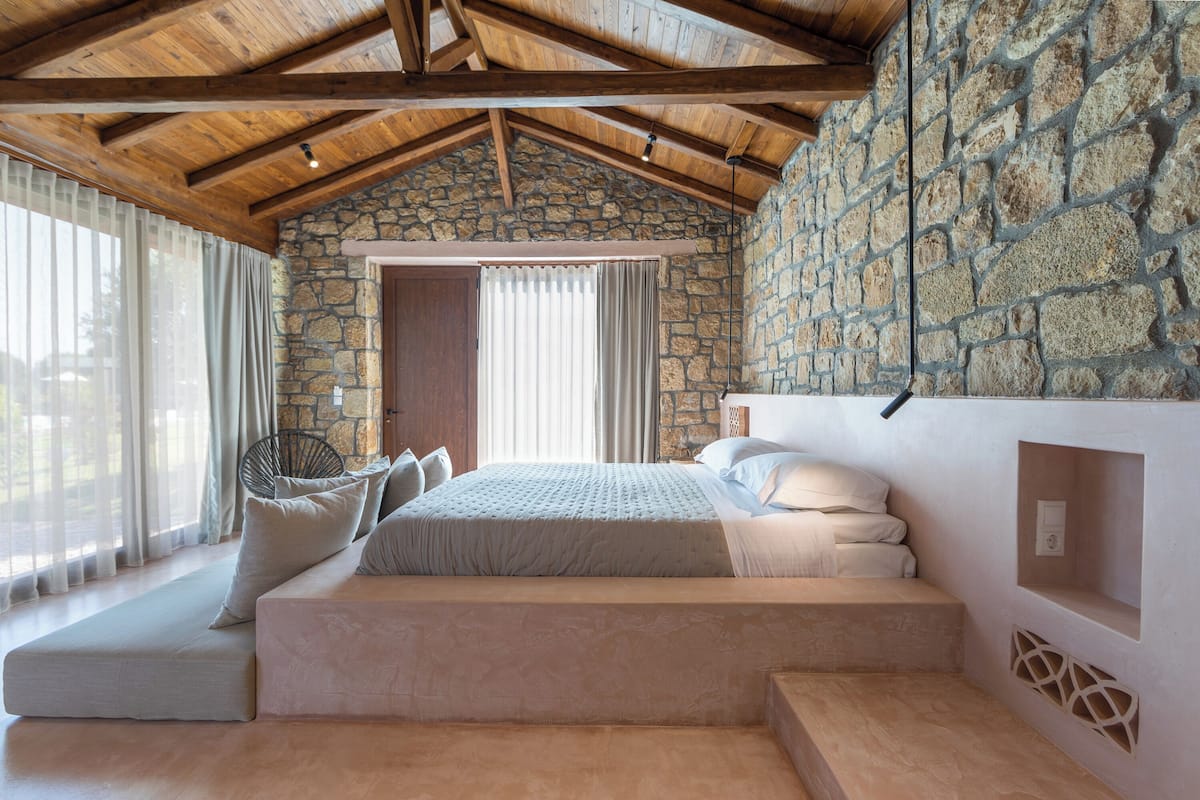 A bedroom with a stone wall, a wooden ceiling, a bed with pillows, and a window with curtains.