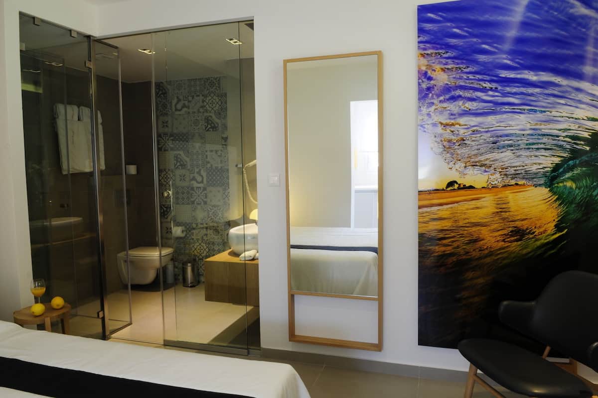 A modern hotel room with a glass shower enclosure, a bed, a large mirror, and a painting of a wave.