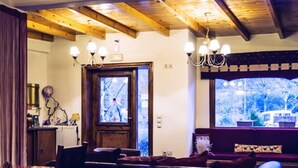 A cozy room with wooden beams, a fireplace, and a view of trees through a window. - Guesthouse Teloni (Gortynia)