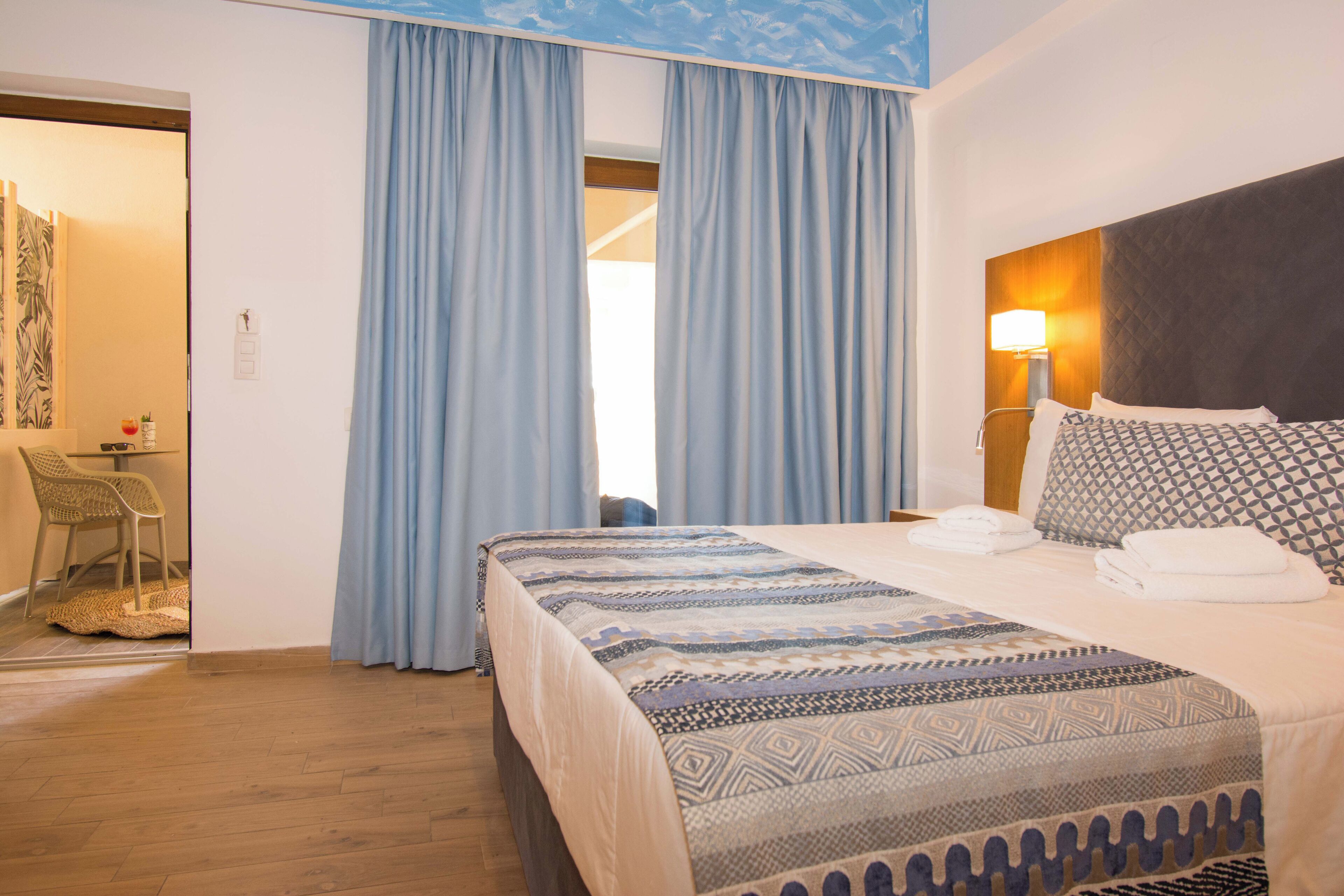 Een hotelkamer met een bed, nachtkastje, lamp en uitzicht op het exterieur door een raam met gordijnen.