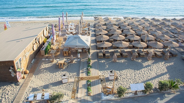 Un complexe hôtelier en bord de mer doté d’une terrasse en bois, de chaises longues, de parasols et d’une enseigne indiquant « Carave ».
