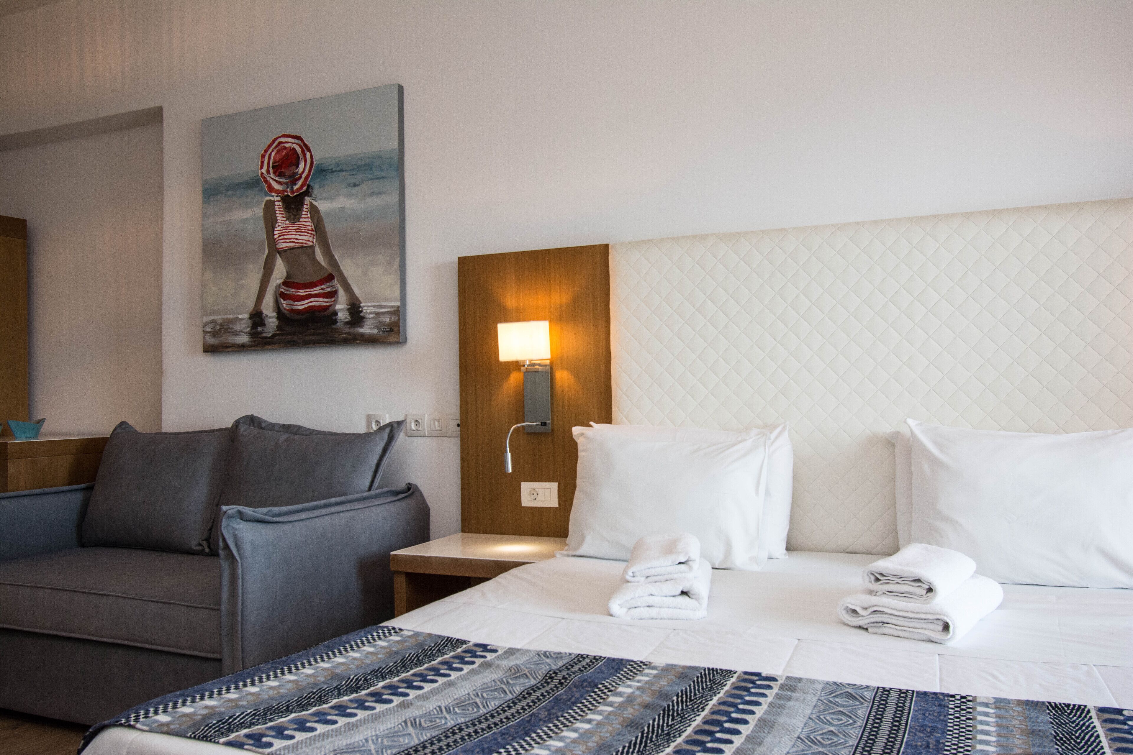 A hotel room with a bed, a nightstand, a lamp, a painting, a sofa, and a view of the sea.