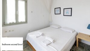 A bedroom with a bed, two framed pictures, a bedside table, and a window with curtains. - Maisons a la Plage (Sifnos)