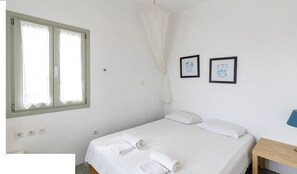 A bedroom with a bed, two framed pictures, a bedside table, and a window with curtains. - Maisons a la Plage (Sifnos)