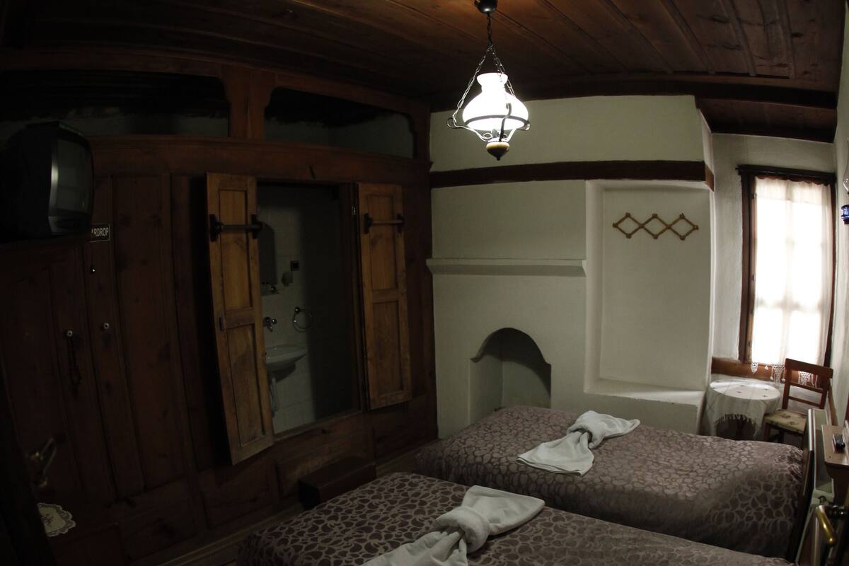Ein Zimmer mit zwei Betten, Holzboden, einer Hängelampe, einem Fenster mit Vorhängen und einem kleinen Tisch mit Stuhl.