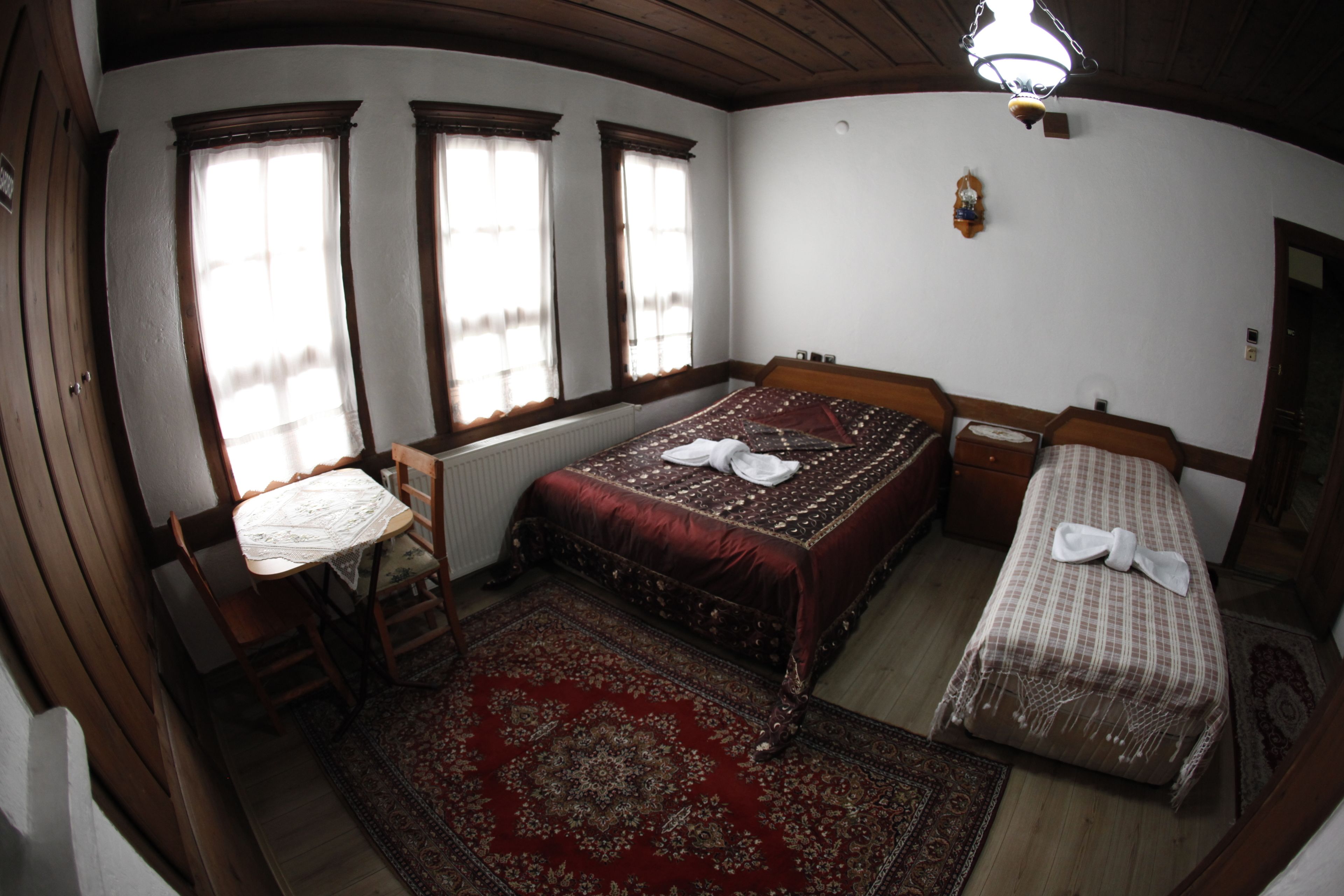 A room with a large bed, a smaller bed, a table, and a chair. There are two windows and a radiator.