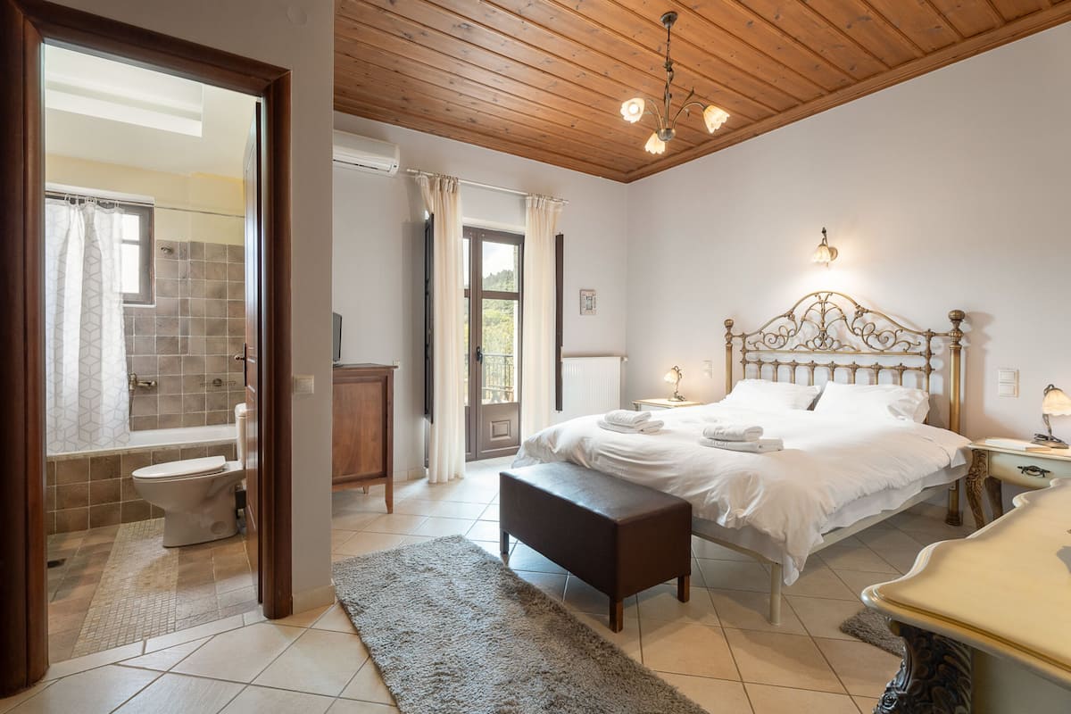 Quarto com teto de madeira, cama com roupa de cama branca, mesa de cabeceira e banheiro com chuveiro e vaso sanitário.