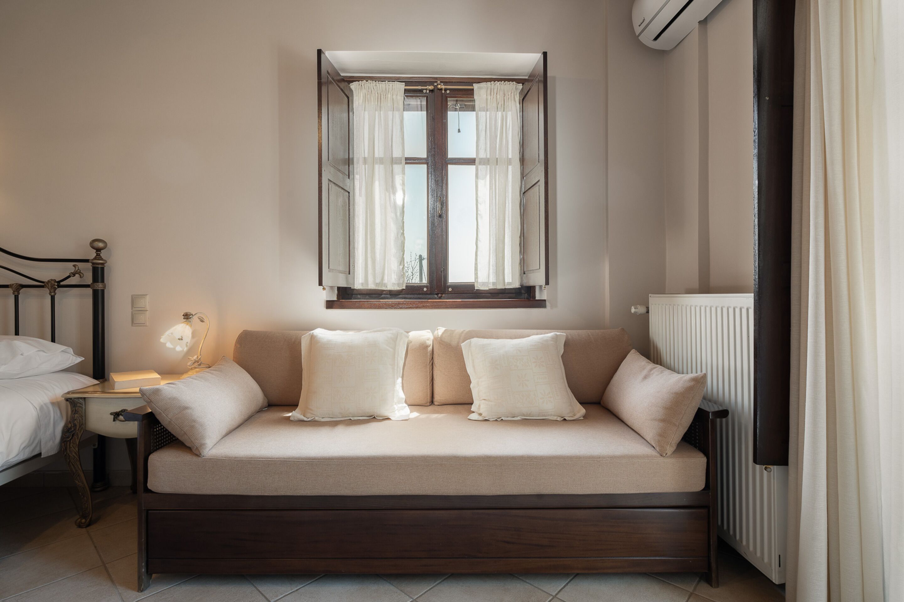 Ein gemütliches Schlafzimmer mit Bett, Nachttisch mit Lampe, einer Couch mit Kissen, einem Fenster mit Vorhängen und einem Heizkörper.