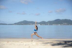 A person performing a yoga pose on a beach with a clear sky and mountains in the background. - My Beach Resort Phuket (Wichit)