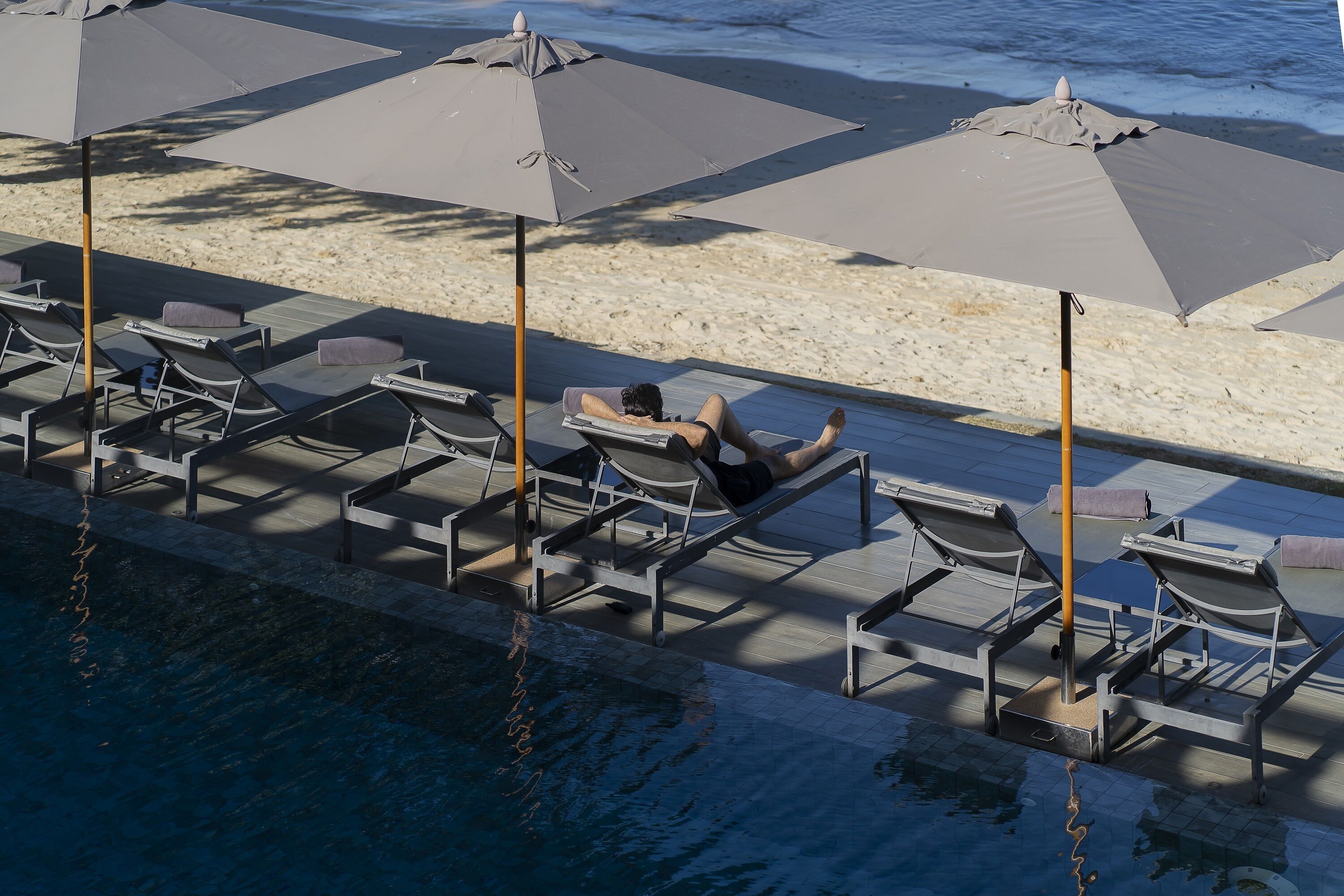 A poolside area with lounge chairs and umbrellas, a pool, and a sandy beach in the background.
