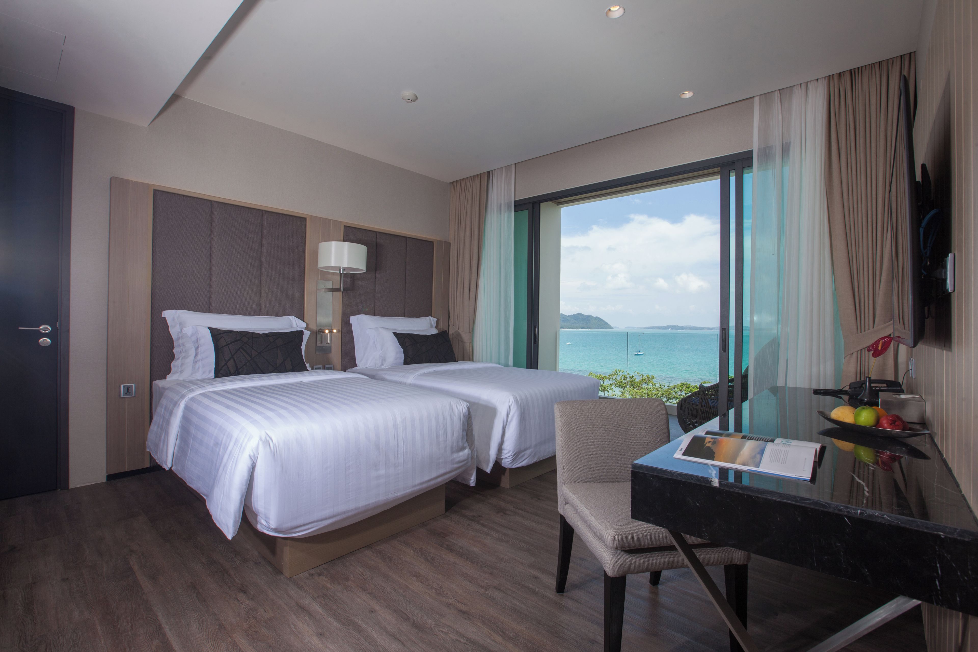 Habitación de hotel con dos camas, un escritorio con silla, un ventanal con vista al mar y un televisor de pantalla plana montado en la pared.