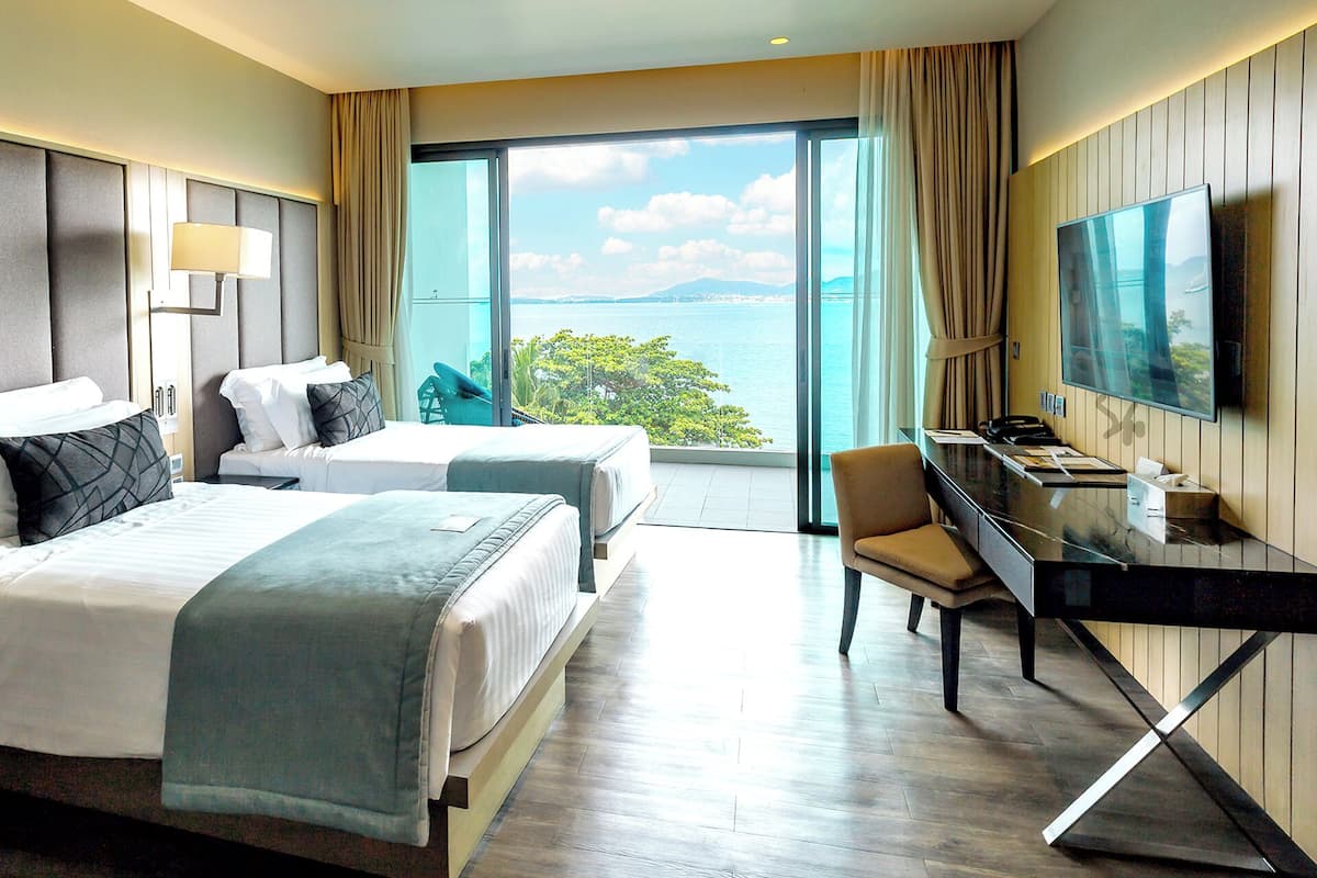 Une chambre d’hôtel avec deux lits, un bureau, une chaise, une télévision et une grande fenêtre donnant sur la mer.