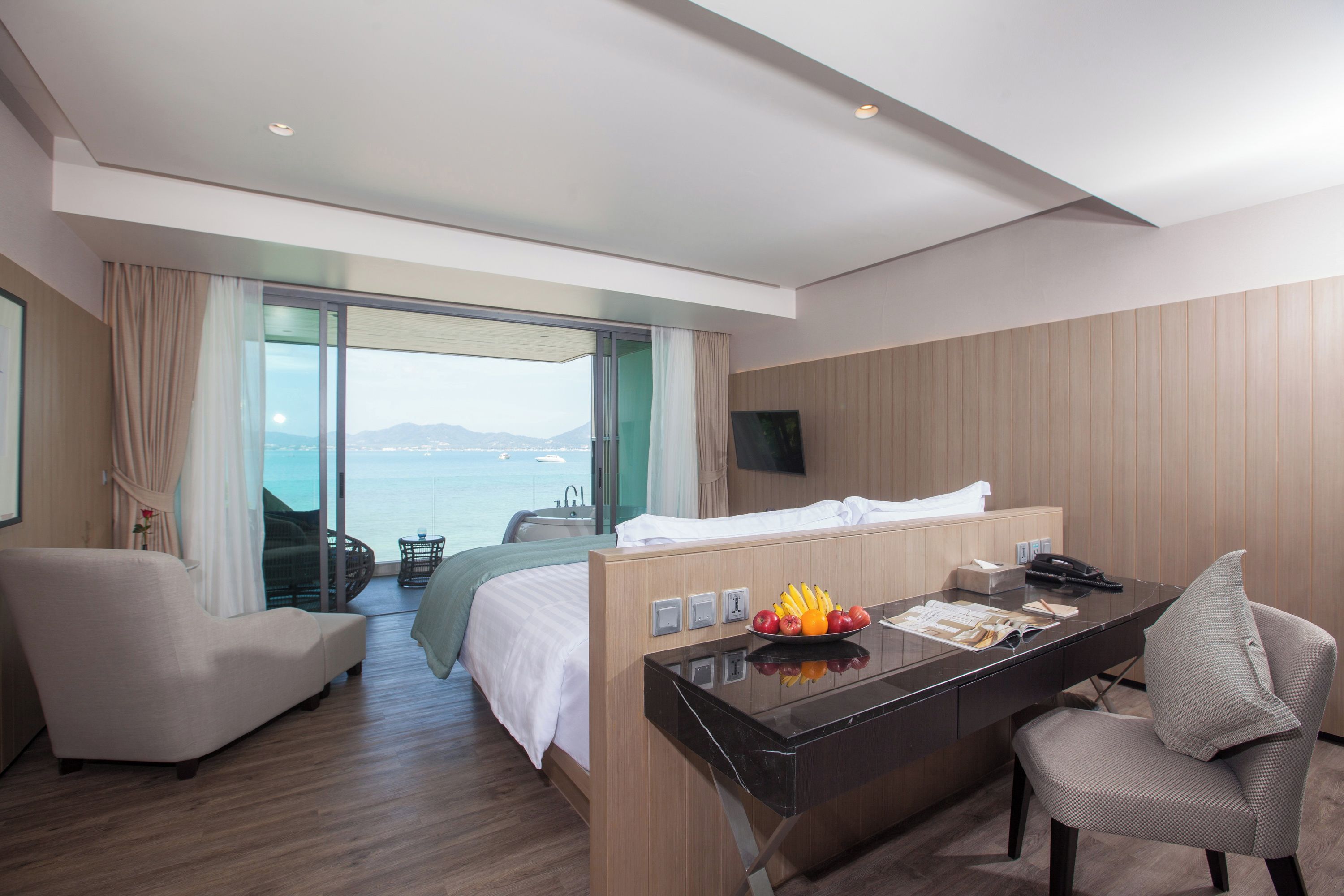 A hotel room with a bed, a desk, a chair, a TV, and a view of the sea.