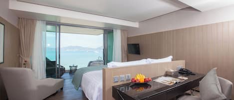 A hotel room with a bed, a desk, a chair, a TV, and a view of the sea.