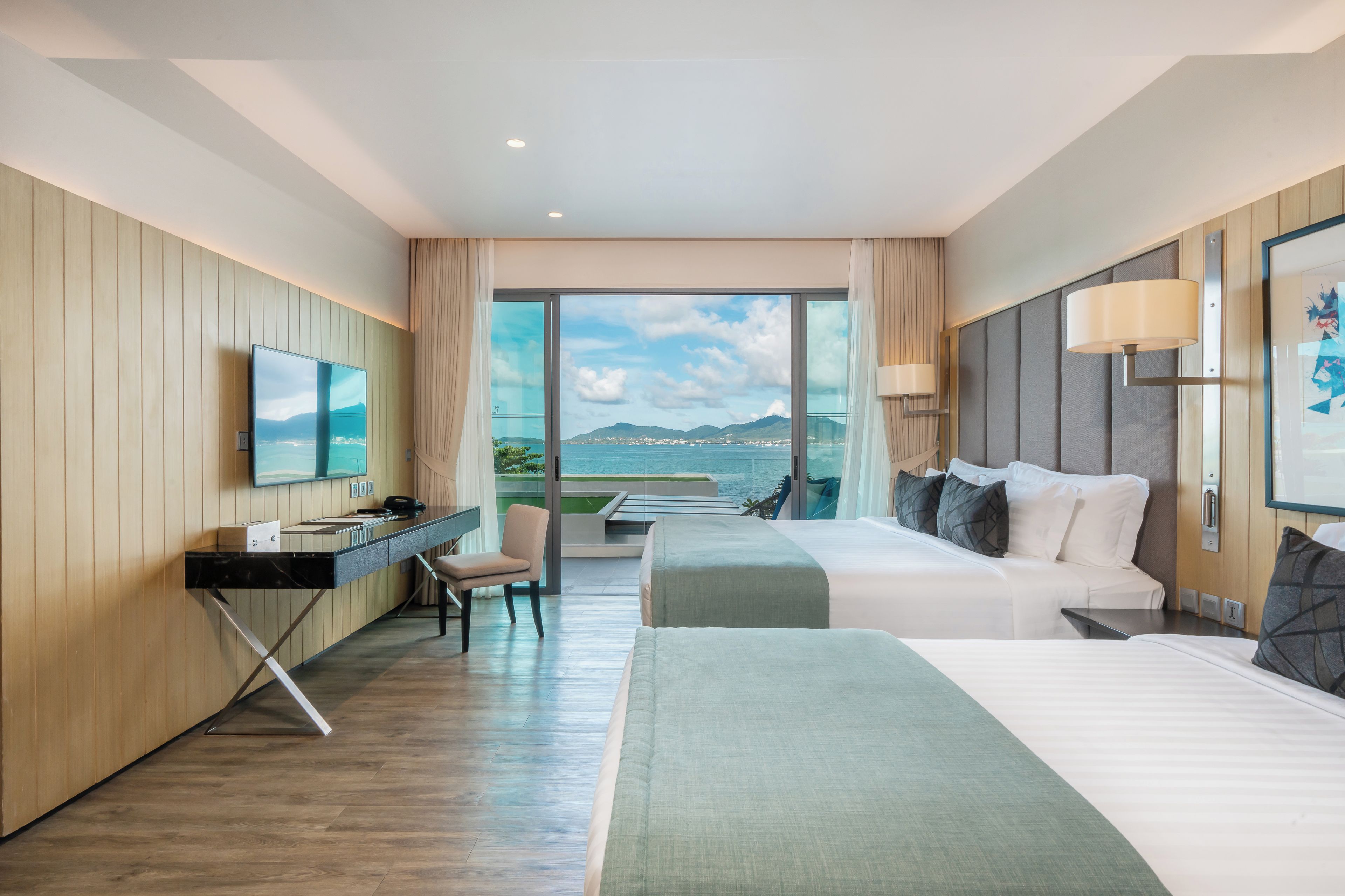 A hotel room with a bed, a desk, a chair, a TV, and a large window with a view of the sea.