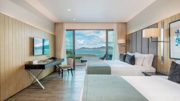 A hotel room with a bed, a desk, a chair, a TV, and a large window with a view of the sea.