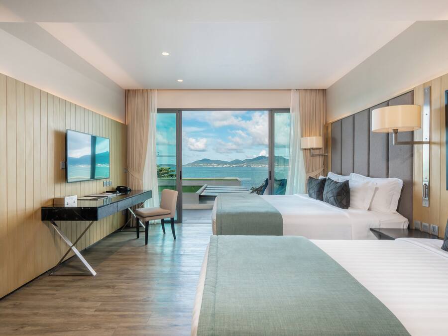 A hotel room with a bed, a desk, a chair, a TV, and a large window with a view of the sea.