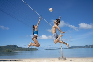 Dos personas jugando voleibol de playa en una arena con un cielo despejado y agua de fondo.