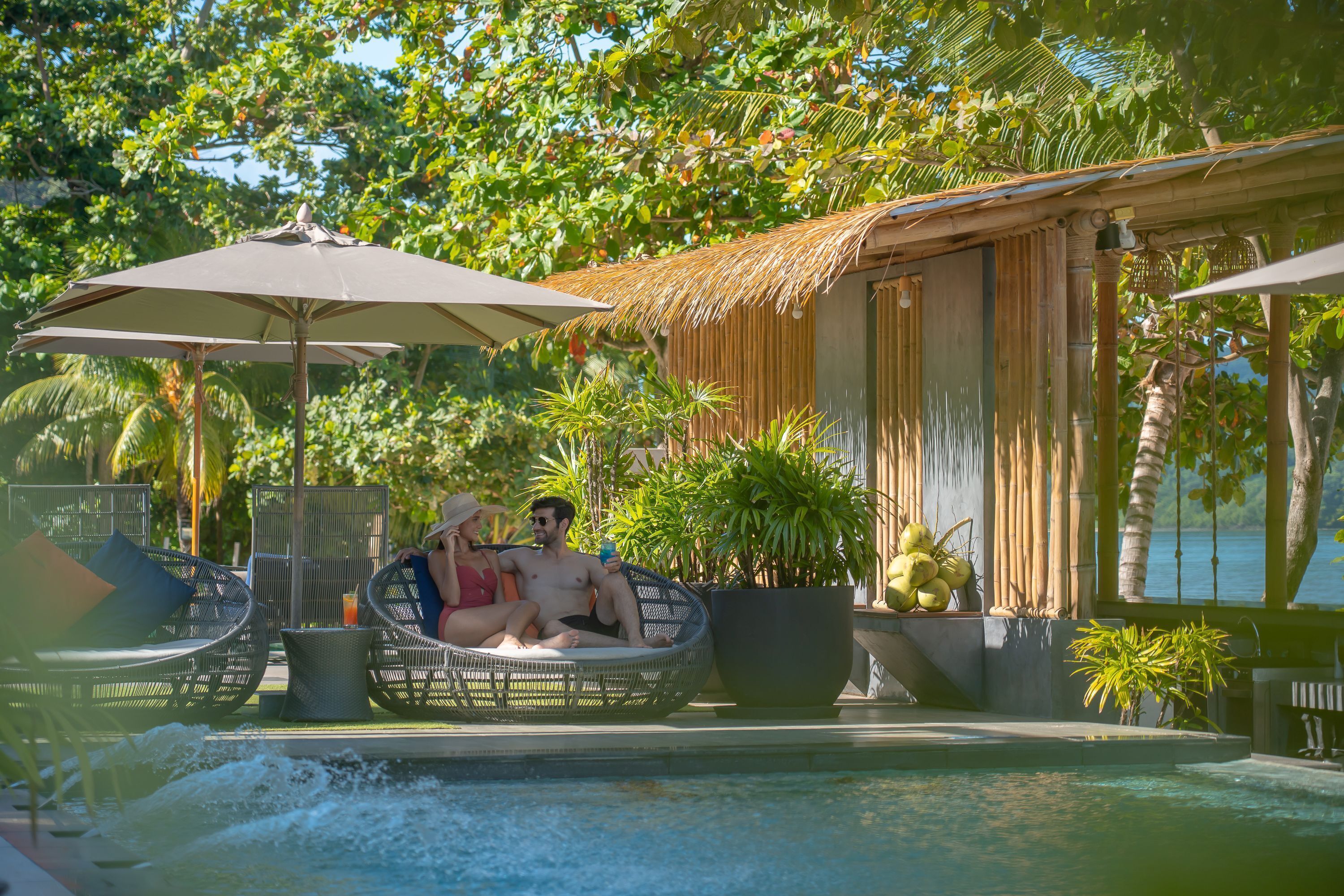 A couple relaxing on a wicker lounge chair by a pool, with umbrellas and tropical plants in the background.