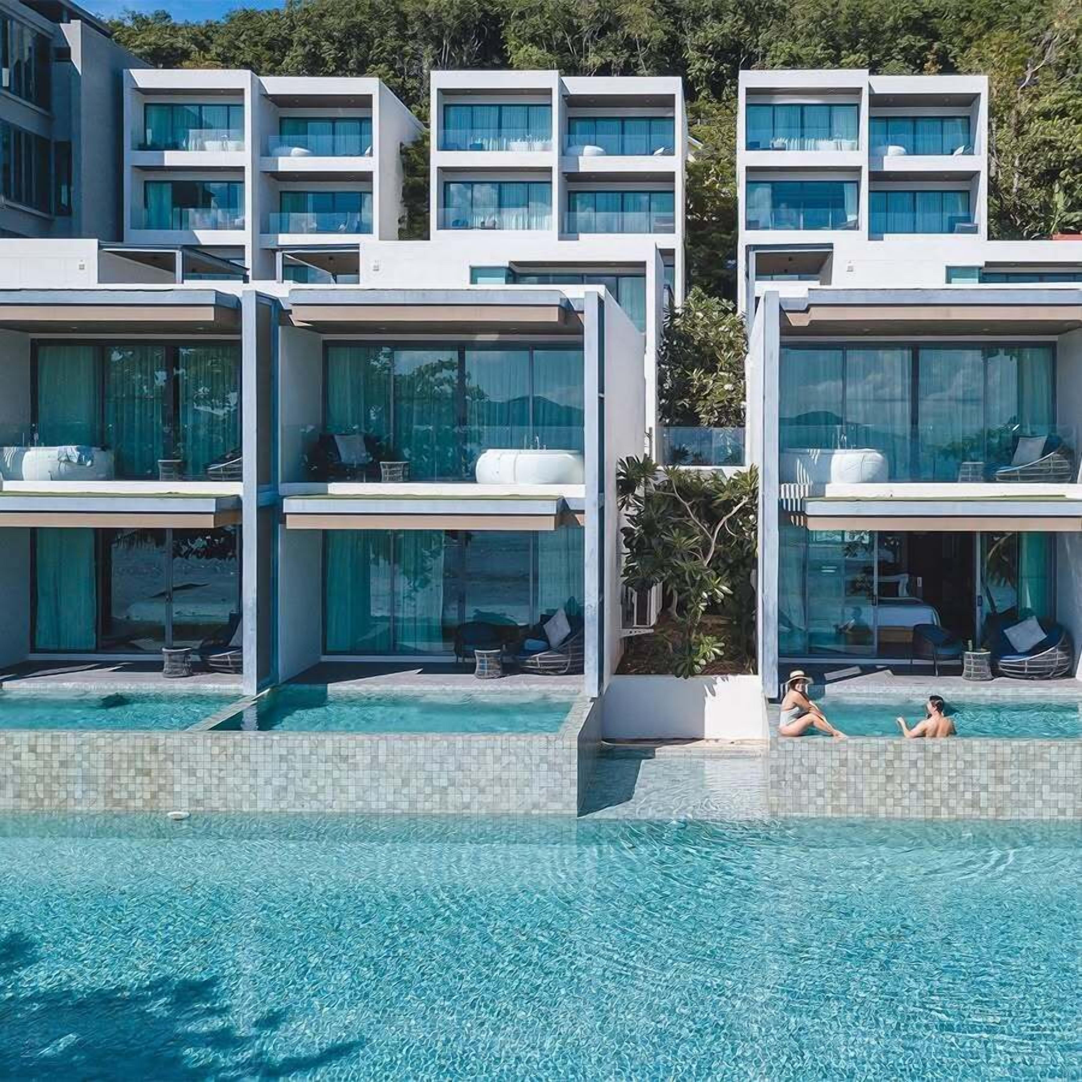 Un hotel moderno con zona de piscina, que cuenta con varios balcones con barandillas de vidrio y mobiliario exterior.