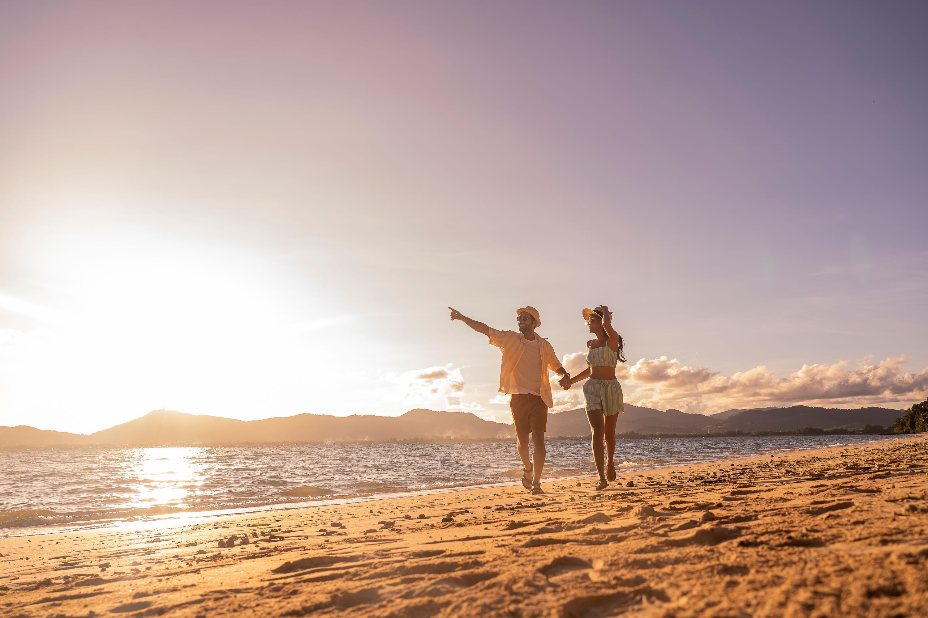 A beach scene with two people walking on the sand, holding hands, and facing the ocean.