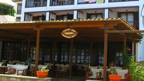 Un edificio de varios pisos con balcones, una cafetería llamada "Maro" y mesas y sillas para disfrutar al aire libre.