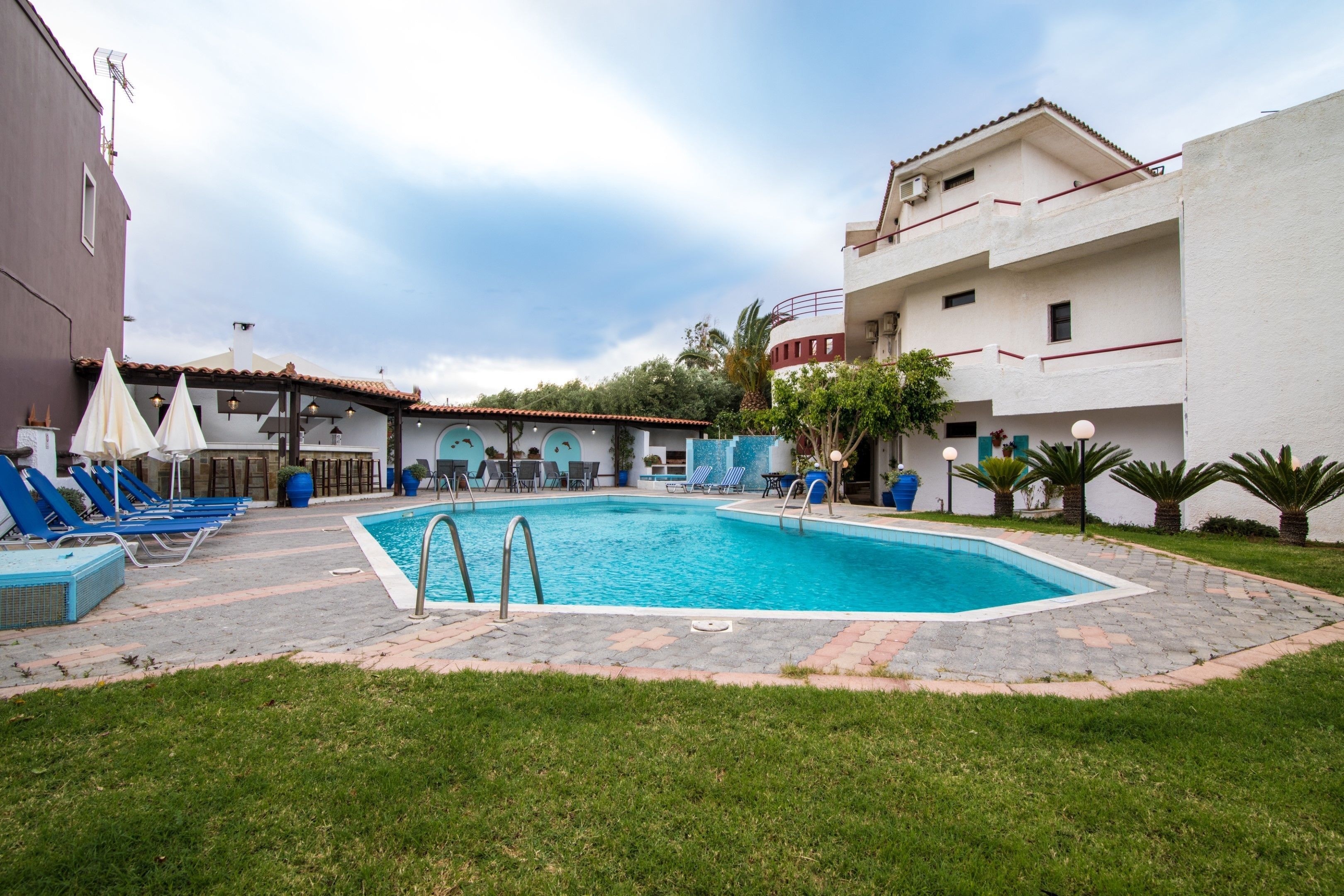 A swimming pool with lounge chairs and umbrellas, surrounded by buildings and greenery.
