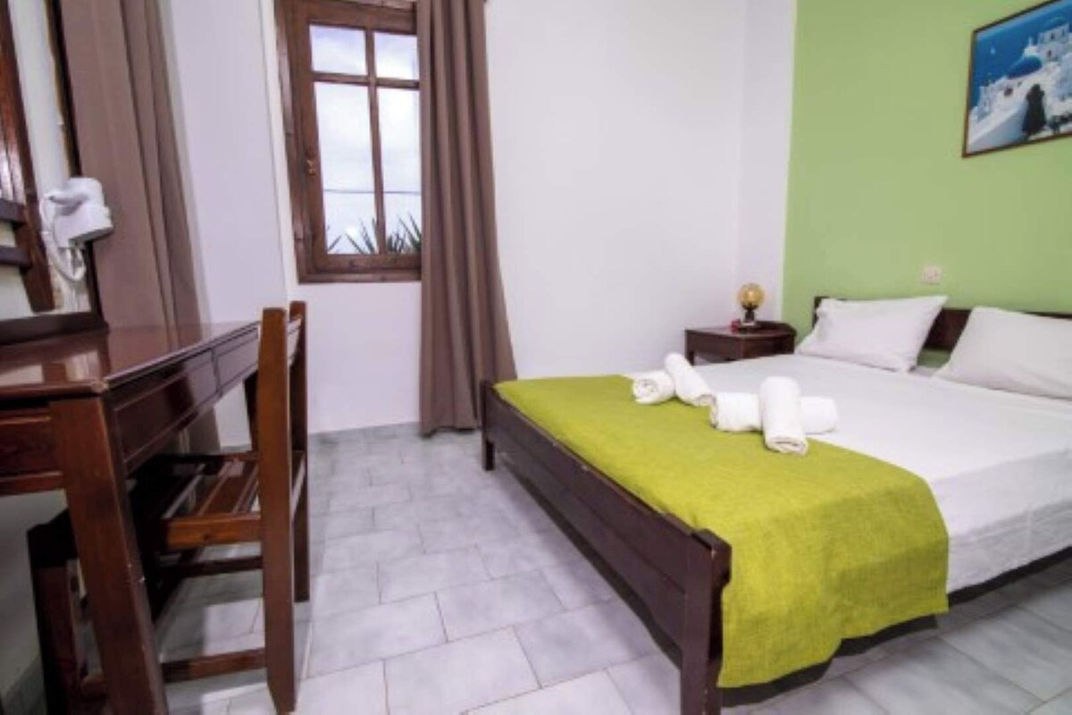 A hotel room with a bed, a desk, a chair, a nightstand, and a window with curtains.